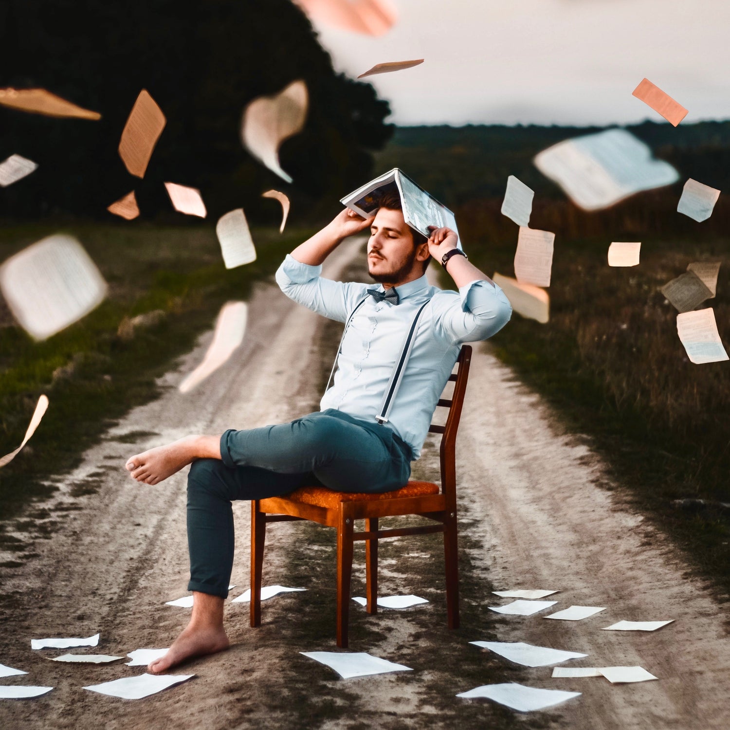 Man sitting in chair in the middle of a road with a book protecting his head from all the papers tumbling down around him