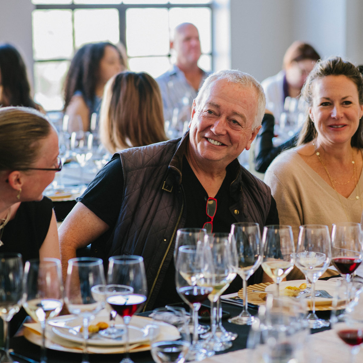 A group of people seated at a dining table filled with wine glasses, plates, and food in a bright indoor event space.