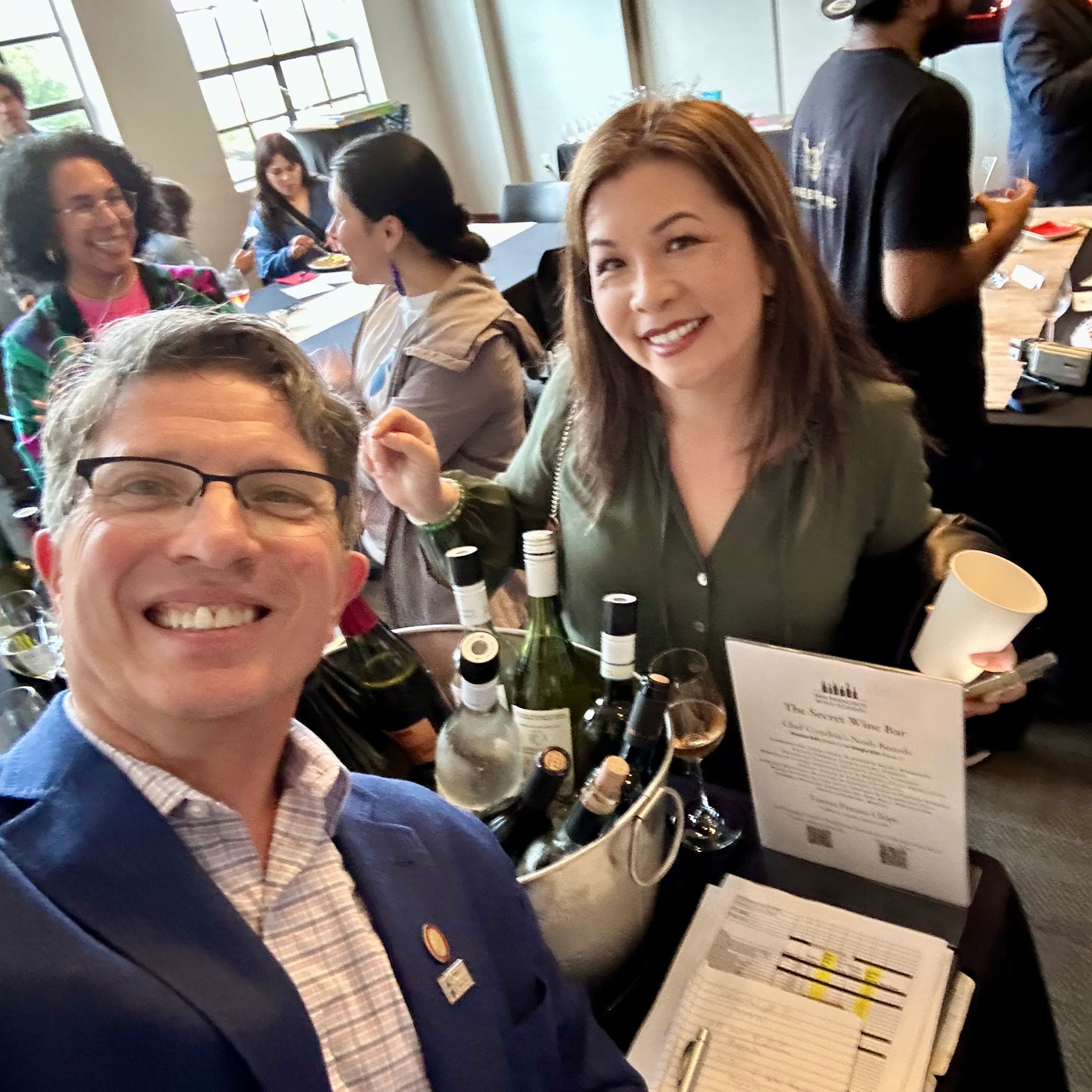 Two people posing for a selfie at The Secret Wine Bar at San Francisco Wine School with wine bottles and a table and other guests in the background.