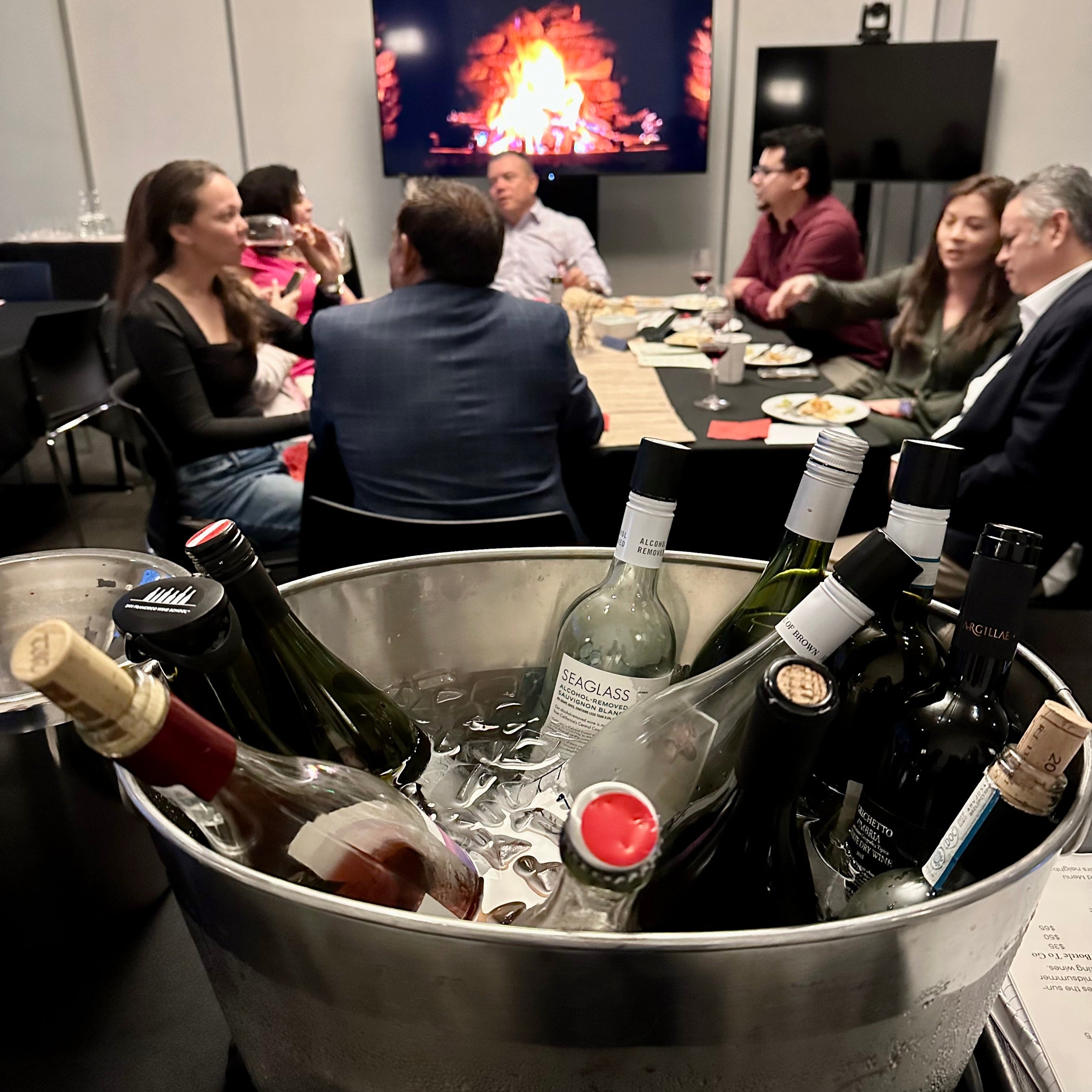 People sitting around a table with wine bottles in an ice bucket in the foreground with a video screen displaying a fire in a fireplace in the background at the Secret Wine Bar in South San Francisco