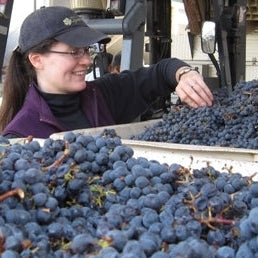 A smiling woman wearing a dark cap and glasses reaches toward a large pile of dark purple grapes in a winery setting, with a forklift visible in the background.