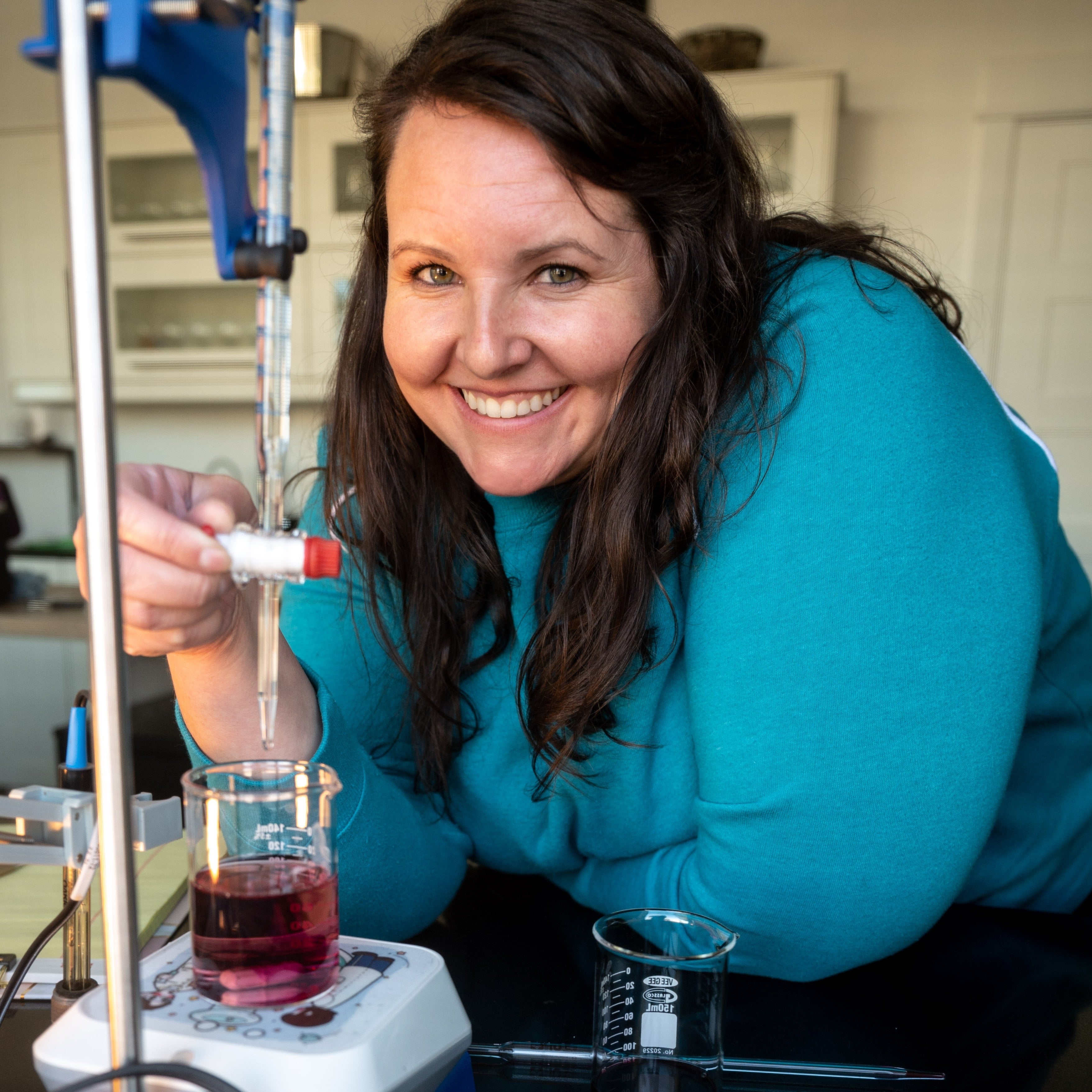 A smiling woman with long dark hair wearing a teal sweatshirt leans over a lab bench, holding a pipette above a beaker containing a dark red wine. Laboratory equipment, including a blue clamp stand and a white magnetic stirrer, are visible in winery lab setting.