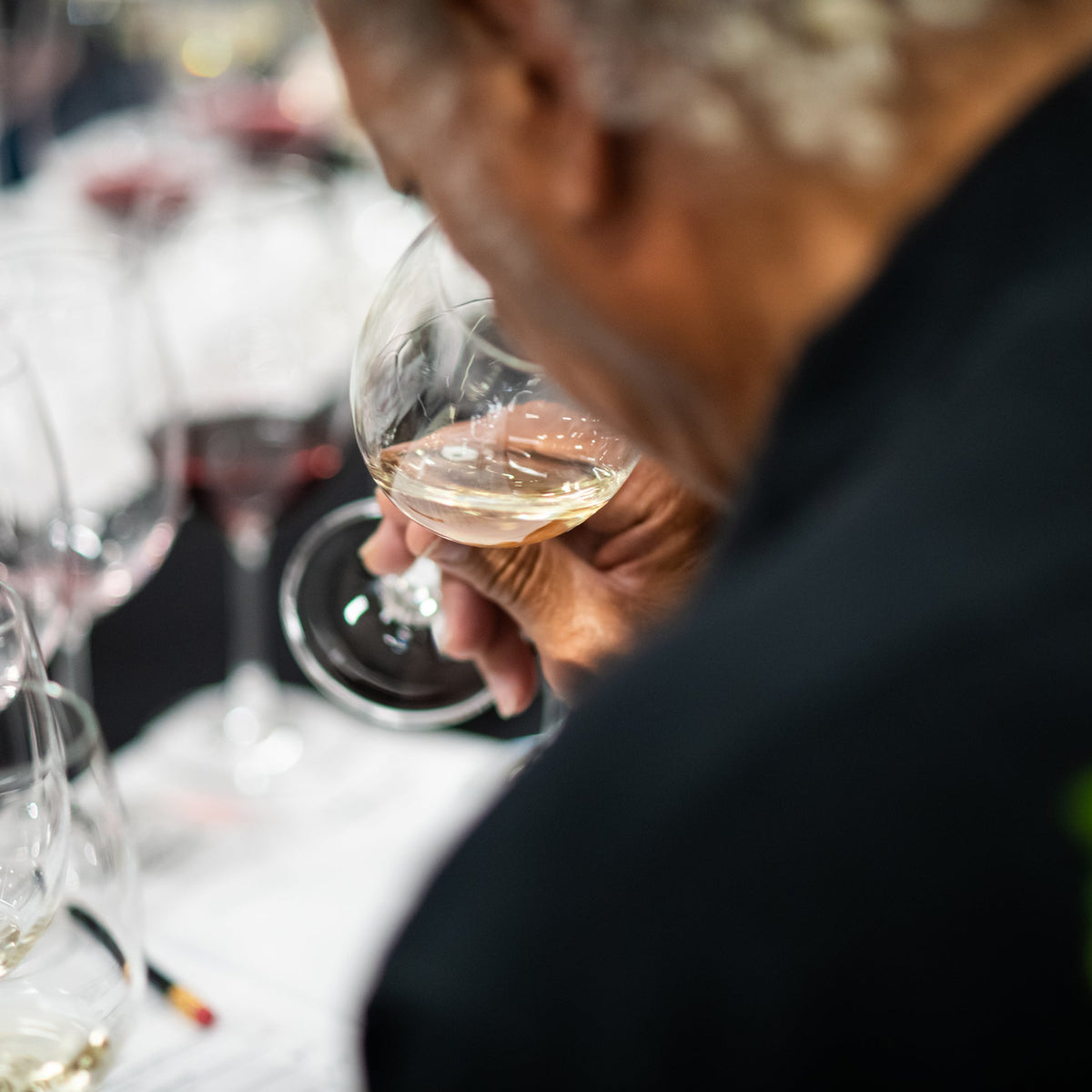 A close-up lifestyle shot of a person holding a glass of white wine at a wine tasting event, with other wine glasses and a blurred background.