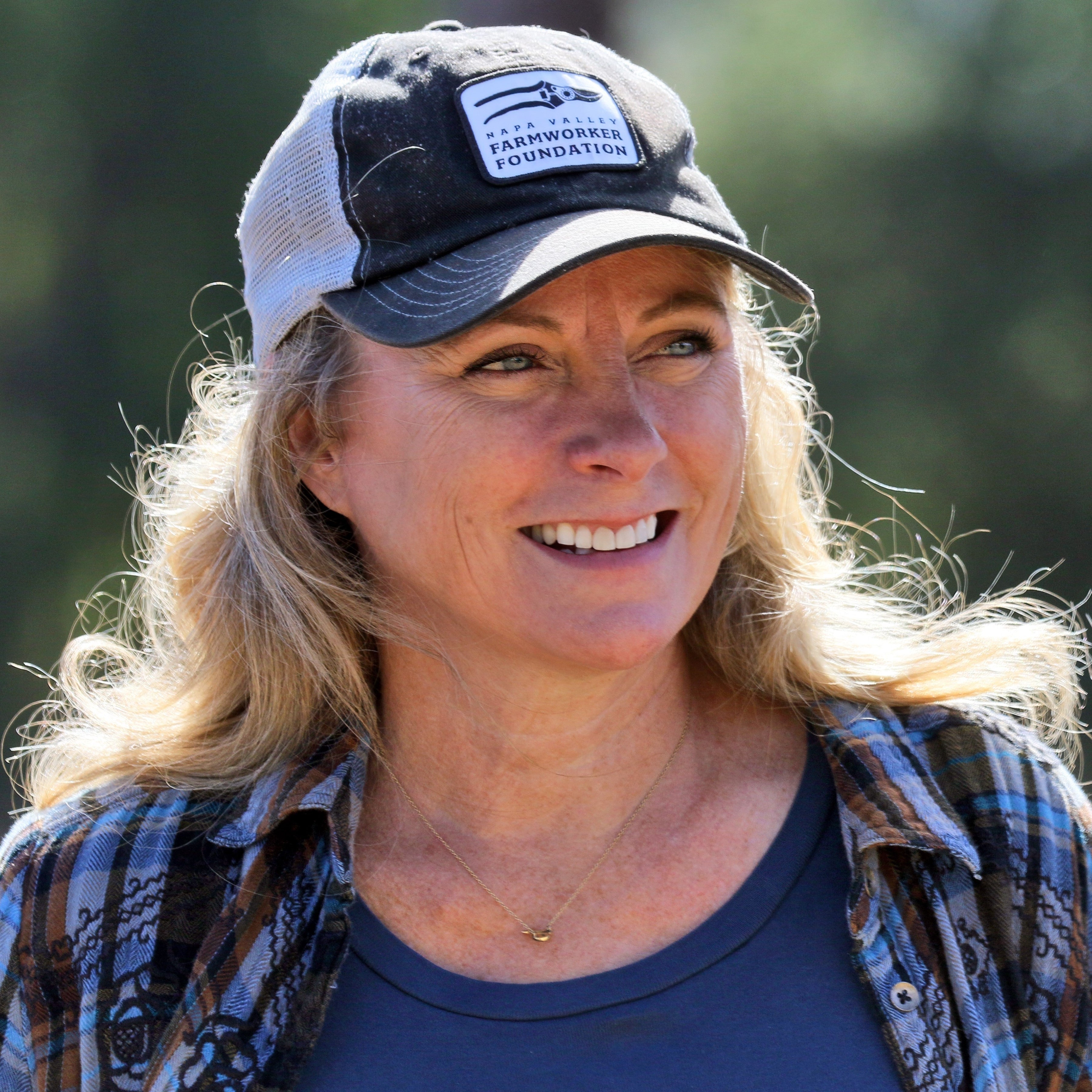 A smiling blonde woman named Debra Becker Lamb who is the Owner of Lost Valley Wines.  She appears outdoors wearing a dark trucker hat with a 'FARMSHOPES AGRICULTURAL FOUNDATION' patch, a blue patterned flannel shirt, and a navy top.