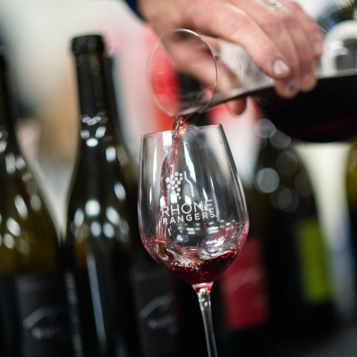 A close-up of a hand pouring red wine from a glass decanter into a Rhone Ranger wine glass, with several wine bottles blurred in the background.