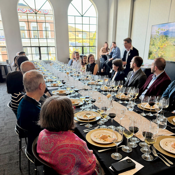 A long banquet table set with black tablecloths, gold plates, wine glasses, and chairs, surrounded by people seated and standing in a large indoor event space with tall arched windows.