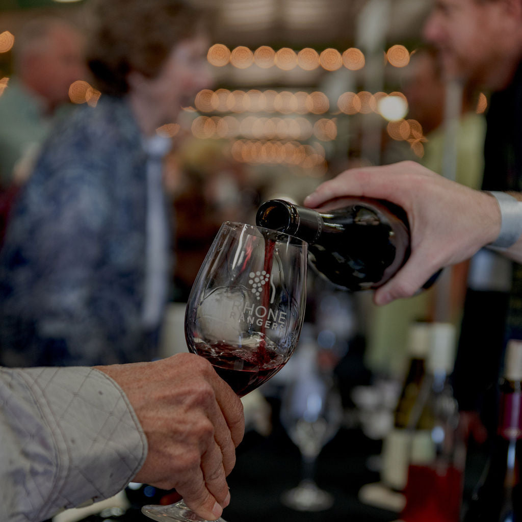 Red wine being poured from a dark bottle into a stemmed Rhone Rangers wine glass held by a person at what appears to be a wine tasting or event. The background is softly blurred with warm lights and other bottles and people.