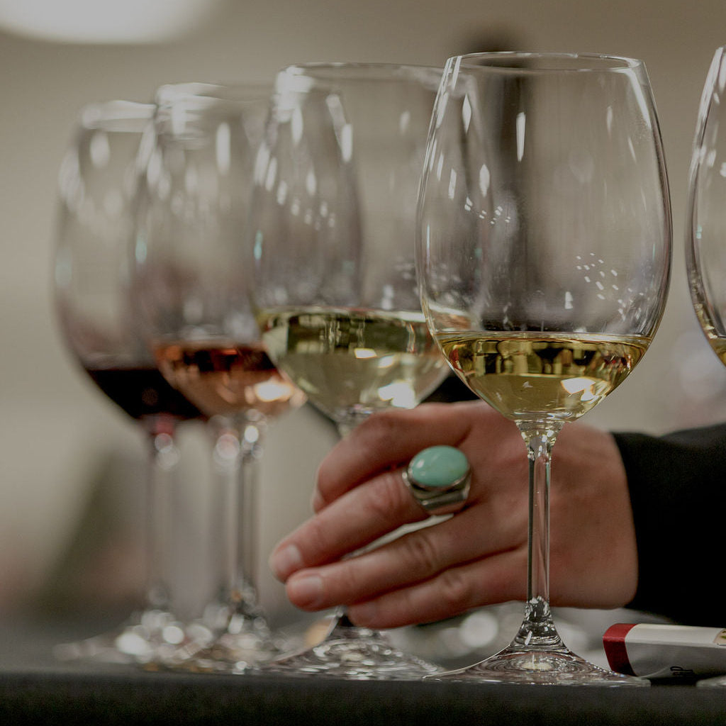 A close-up of a hand wearing a turquoise ring, resting near a row of wine glasses containing red, rosé, and white wine, with a blurred indoor background. 