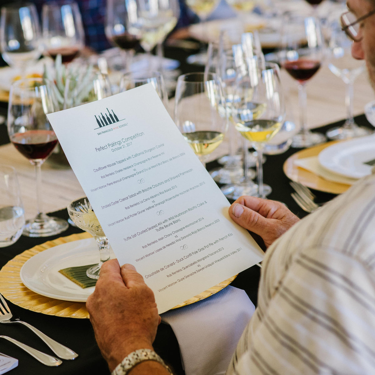 A man seated at a dinner table holds a printed wine pairing menu card. The table is set with multiple wine glasses, plates, and cutlery, with another person visible in the background.