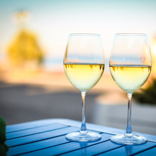 Two glasses of white wine are placed on a blue surface with a blurred background featuring greenery and a body of water. The glasses are filled with light-colored Chardonnay wine, and the scene suggests a serene, outdoor setting, possibly indicative of the type of experience offered by the wine school.