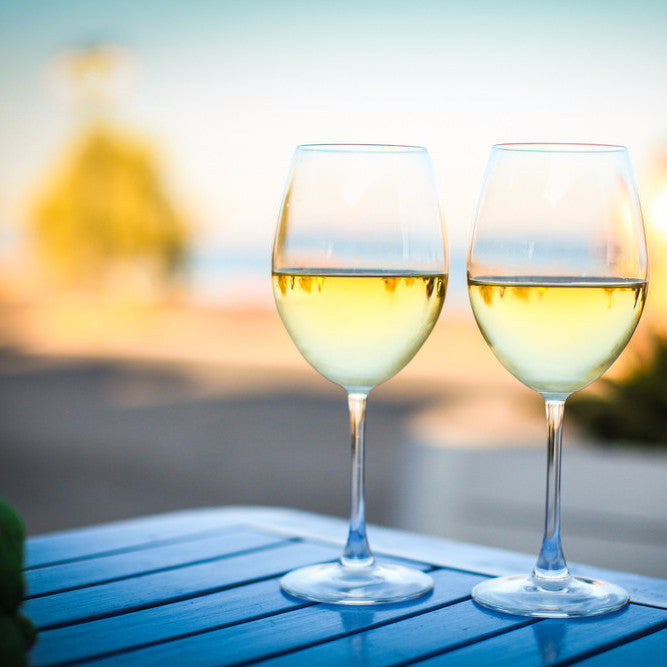 Two glasses of white wine are placed on a blue surface with a blurred background featuring greenery and a body of water. The glasses are filled with light-colored Chardonnay wine, and the scene suggests a serene, outdoor setting, possibly indicative of the type of experience offered by the wine school.