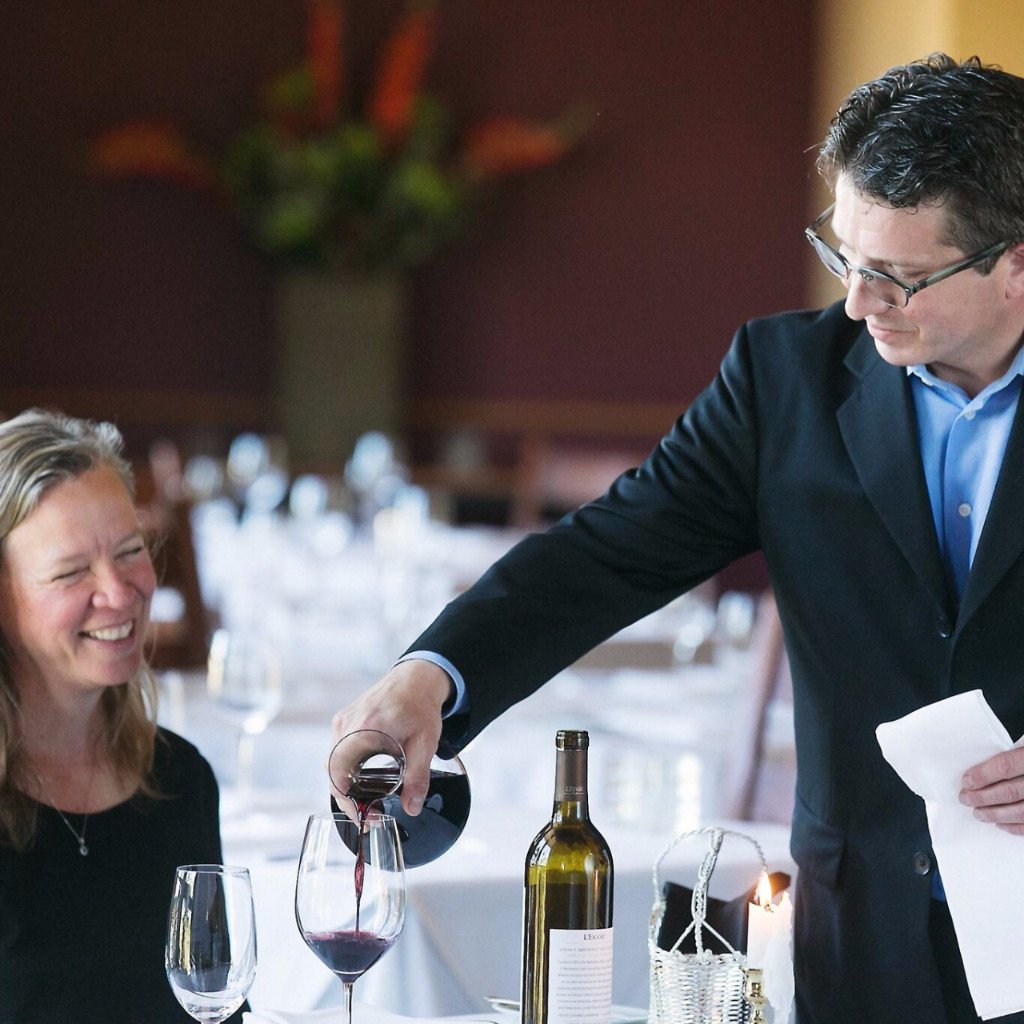 A wine server pouring wine into a glass for a smiling guest at a dining table in a restaurant.