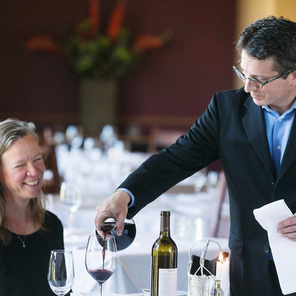 A wine server pouring wine into a glass for a smiling guest at a dining table in a restaurant.
