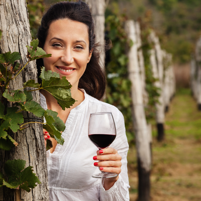 A woman holding a glass of Syrah red wine, standing in a vineyard with grapevines and greenery in the background.